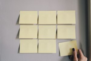 A grey wall with yellow post-it notes stuck in a 3 by 3 grid pattern. A hand is placing the bottom right note into the grid to complete it.
