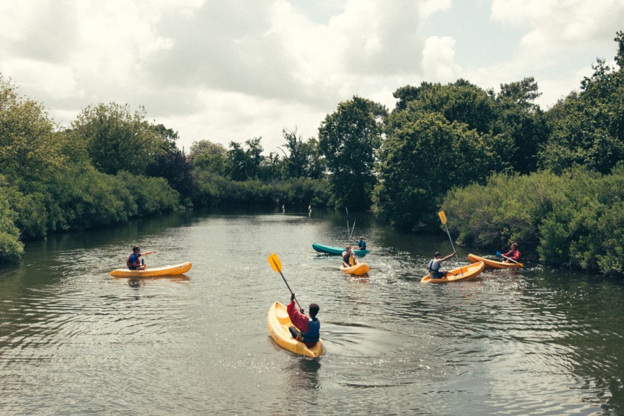 A photo of a kayaking lesson with a teacher and a small group of beginners in a wide, tree-lined river or canal.