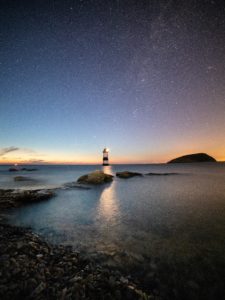 A small lighthouse in the centre of the image warns a rocky shoreline amongst a dusky blue sunset sky with stars visible.