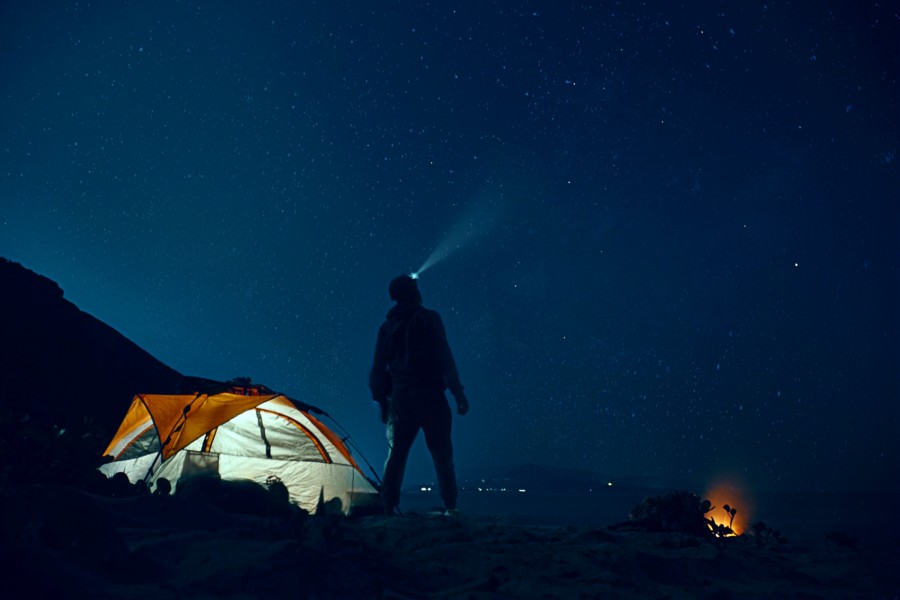 Night-time camping scene with a dark blue sky full of stars and the only light coming from an orange tent to the left. A lone silhouetted star-gazer looks up to the stars with a bright headtorch beaming towards the sky. A glowing campfire is on the right.