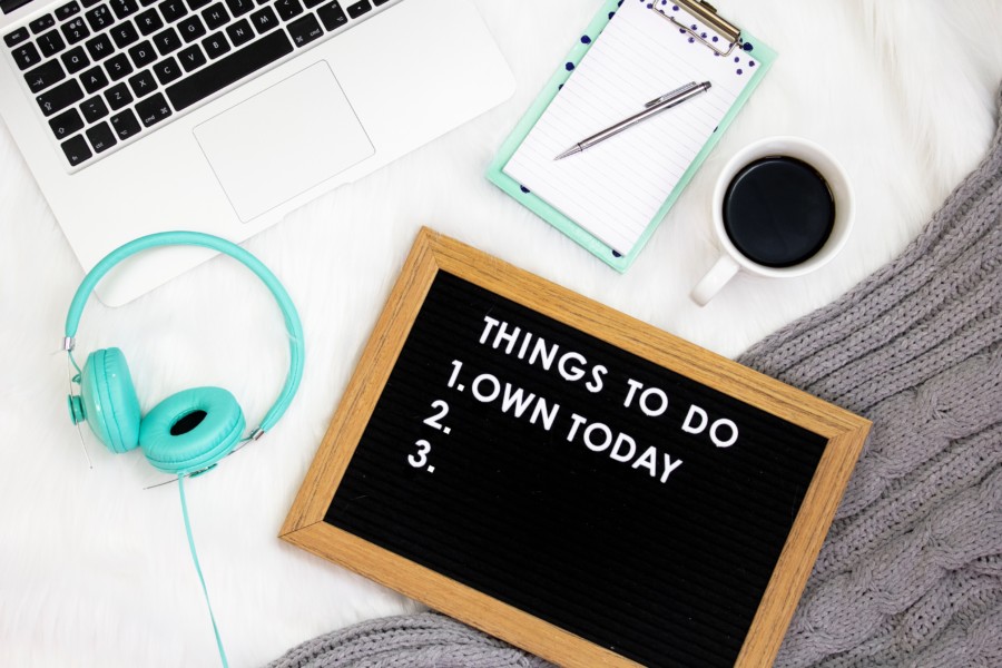 A flatlay photo of a white desk surface displaying a few items. A black rope noticeboard with walnut frame reads: "Things to do: 1. own today. 2. [empty space] 3. [empty space]. A pair of bright neon turquoise wired headphones, a laptop, turquoise notebook, black coffee in white mug and grey cable knit jumper sleeve decorate the image edges.
