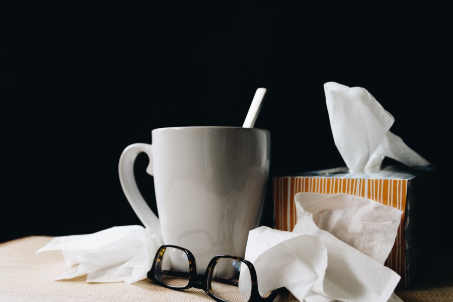 A white mug of a presumably hot drink with a teaspoon sticking out of it is positioned next to an orange striped box of tissues with some of the scrunched up tissues next to it. A pair of up-turned glasses also sit nearby suggesting a dishevelled, ill-health situation.