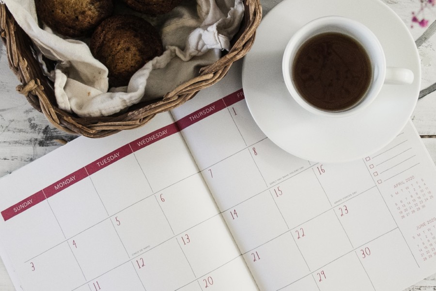 An americano coffee in a white cup and saucer sit on top of a calendar notebook diary, with a wicker basket of baked treats to accompany them.