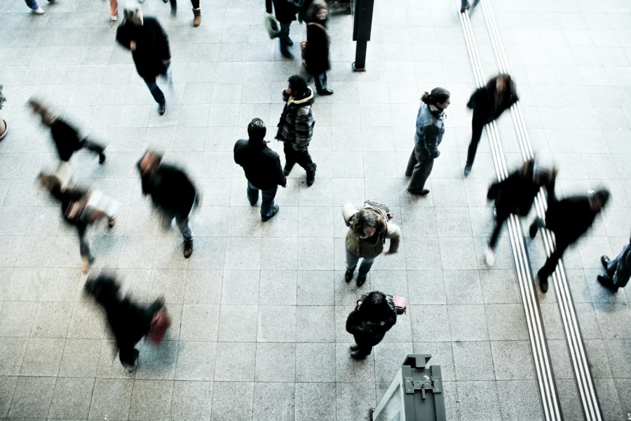 A busy scene of people walking in different directions through an urban square. Some are blurred from the long-exposure effect of the photography.
