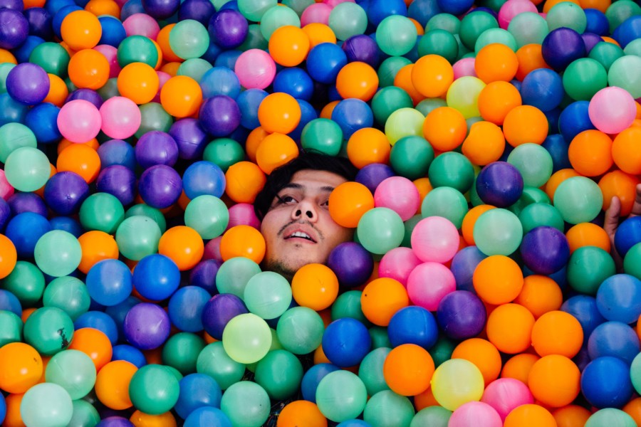 A person seemingly drowns in brightly coloured plastic balls in a ballpit where only their face is visible with a strained facial expression peering through.