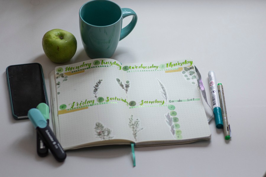 A planning notebook is open with hand drawn days of the week in green pens with a teal green mug and a green apple positioned above on the white table. The green and turquoise pens used sit to the sides.
