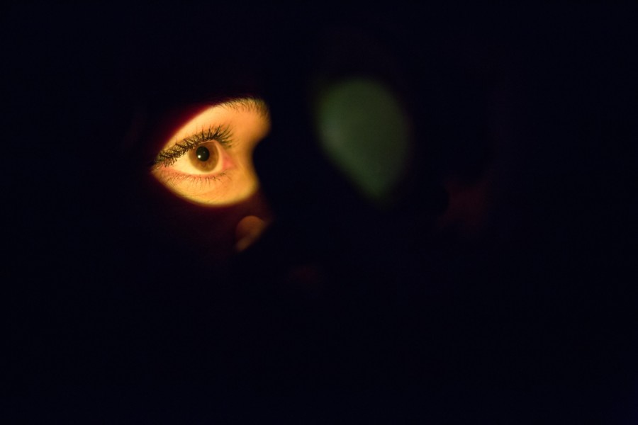 A completely dark room with only a human eye visible in a beam of yellow light projected onto their face. They are looking off into the distant right hand side in thoughtful expression.