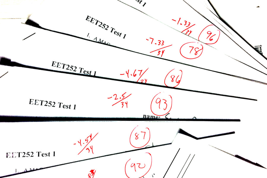 A set of white paper tests that have been graded in red pen with a variety of scores out of 100. The papers are displayed in a fan from the bottom left corner to the top right to just show the titles and the grades on the top but not the content of the test.