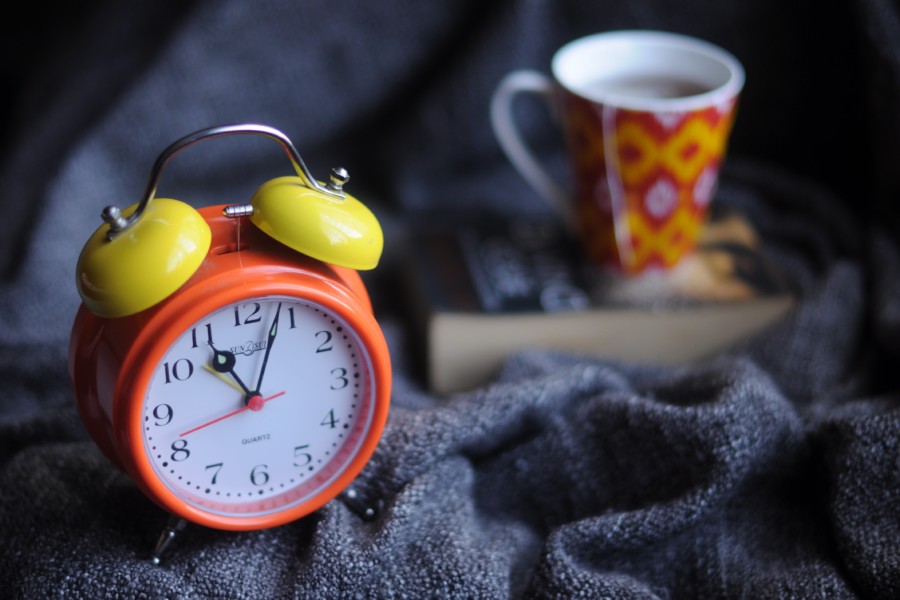 A bright orange and yellow classic analog alarm clock sits on a grey blanket in the focused foreground, while a orange and red patterened mug with a hot beverage inside sits in the blurred background.