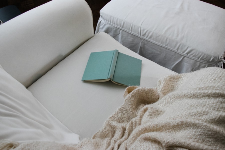 A blue book is open and faces upside down on a white sofa with a beige blanket nearby, indicating a pause in reading it or putting it down because the reader became fed up.