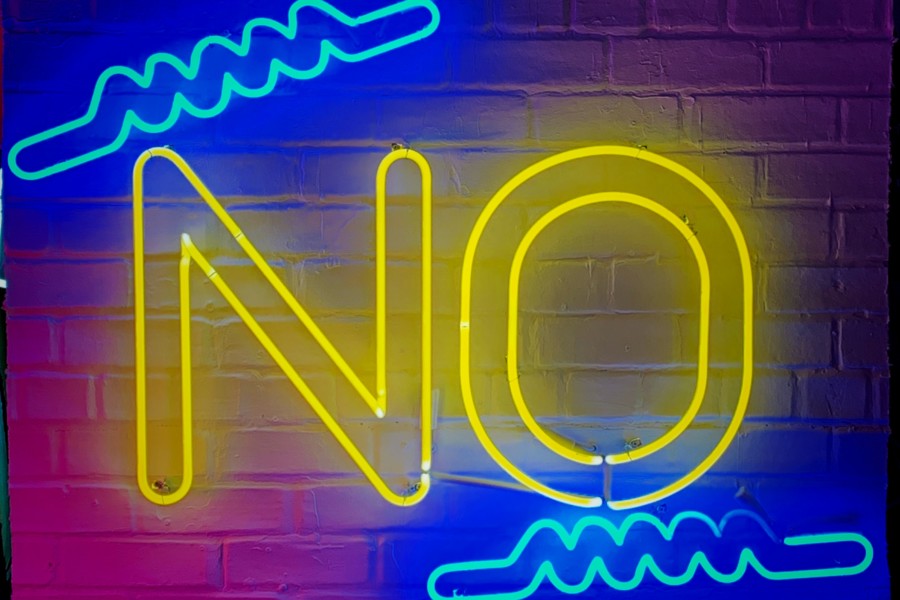 A yellow neon sign spells out the word "NO" in capital letters with some blue squiggly line neon signs decorating and emphasising the No at the top and bottom. A pink tint of light is also on the wall making it a bright lively colour combination and adding some humorous irony to the No