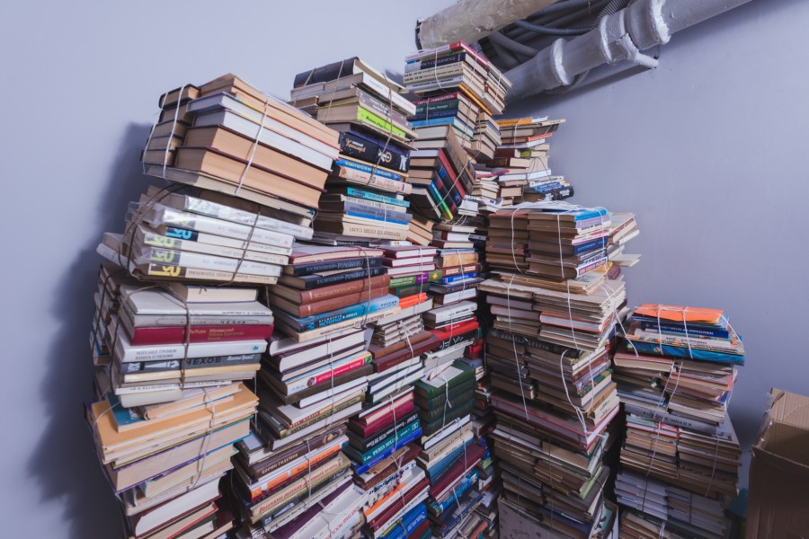 multiple tall yet precariously stacked piles of books against a corner wall, with some books toppling into the next pile in a chaotic scene. Some groups of the books are wrapped together with string however.