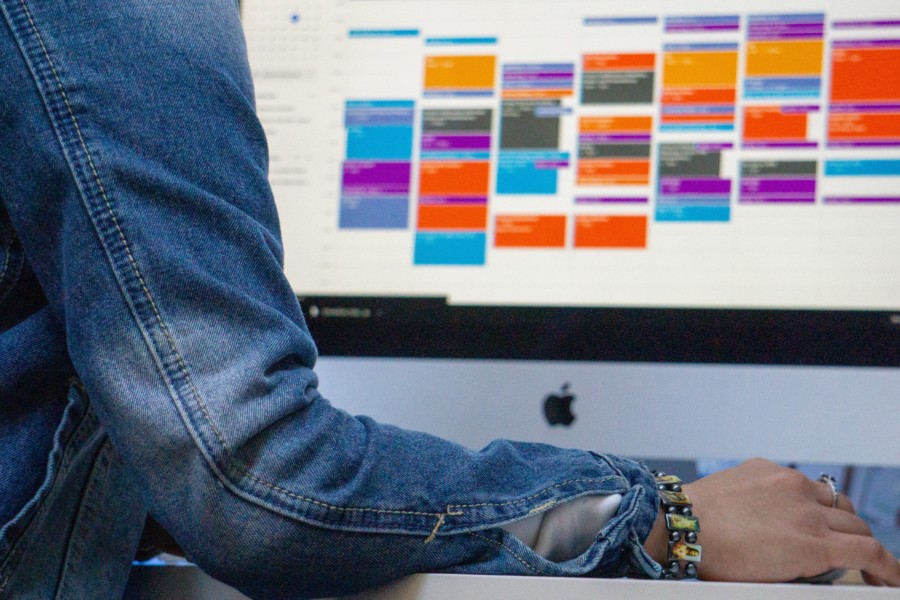 A photo of a person in a blue denim jacket sitting at a mac computer which is displaying an online calendar in week overview. It's showing a very busy layout of intersecting blocks of events on each day that are brightly colour coded.