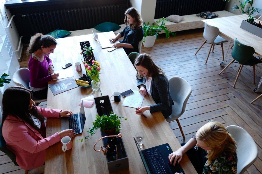 A group of women sitting at a long wooden table co-working together. There's two on one side and 3 on the other in a casual workplace setting with laptops, stationery and mugs around the area.