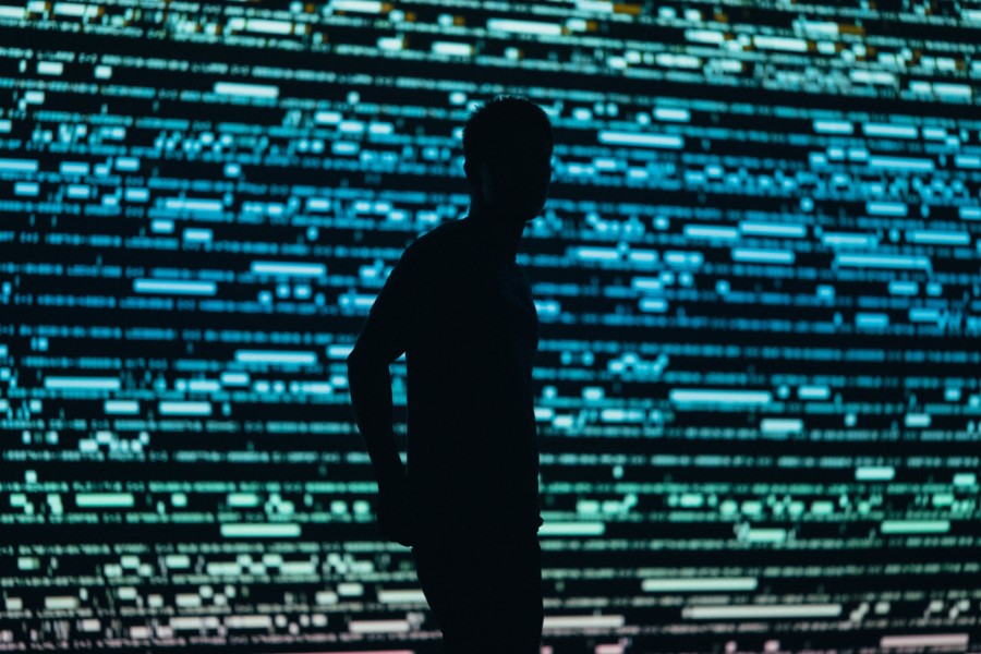 A photo of a person's silhouette stood against the full wall background of electronic blue and green dots and lines that look like computer code or music editing software from afar.