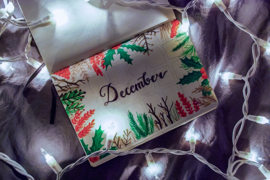 A plain notebook open to reveal the word December handpainted in black paint, along with decorative red and green foliage around the edges of the page. The notebook is on a cosy bed surrounded by fairy lights.