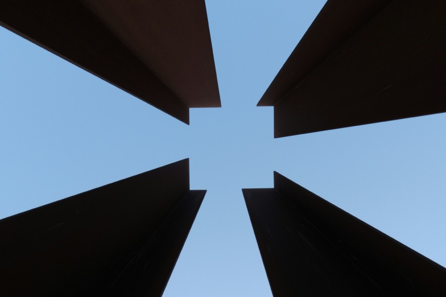 A striking photo taken from beneath 4 angular pillars leaning into the centre of the image from each of the 4 corners, with notches cut from their tips that creates the illusion of a square in the central negative space. The sky behind is pastel blue and the pillars are a dark reddy brown, possibly rusted metal.