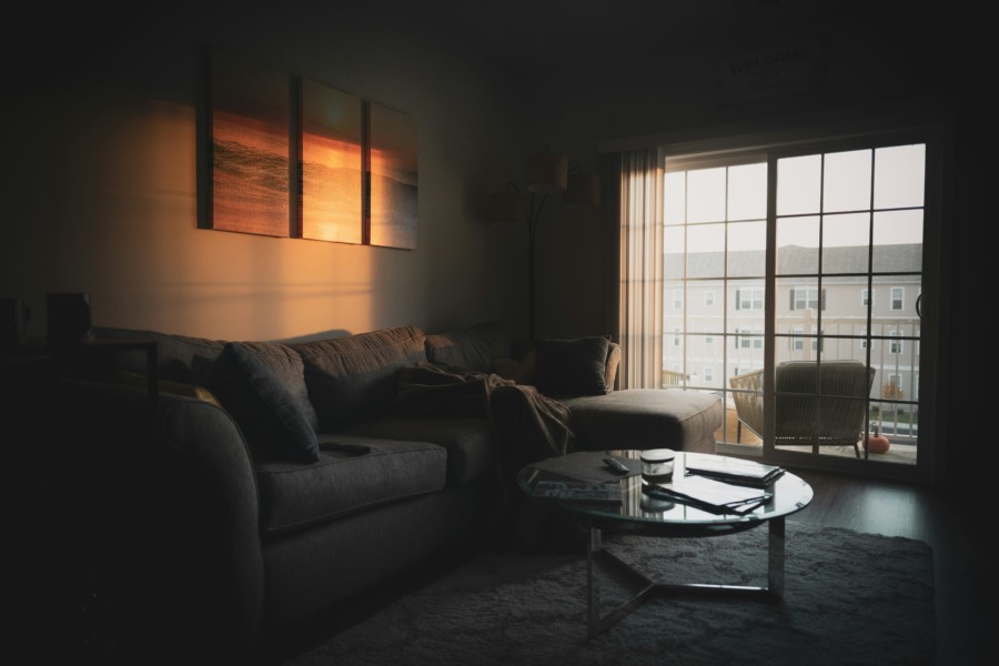 A photo of a dark living room with only a small square of low sunlight illuminating the wall above a well-worn sofa.