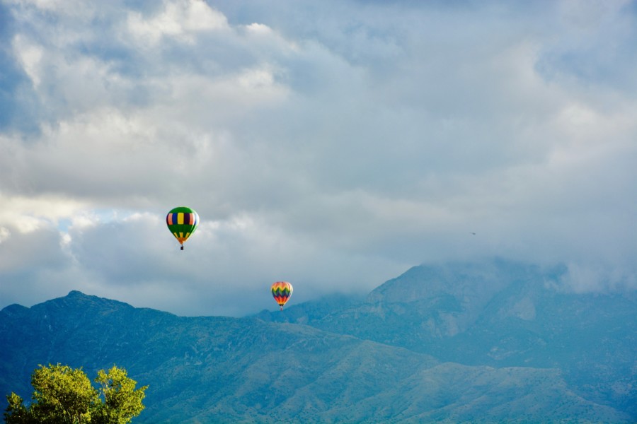 2 hot air balloons ascend majestically across some mountains, their peaks covered in clouds, but the sunshine shining onto the colourful patterned sides of the balloons.