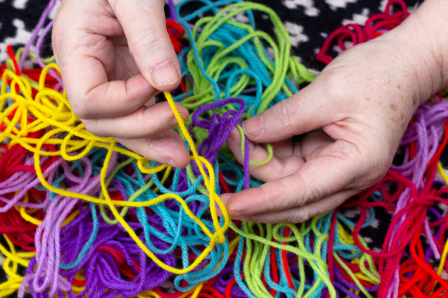 A photo of a person pulling different colourful threads of wool apart above a tangled pile of different colours.